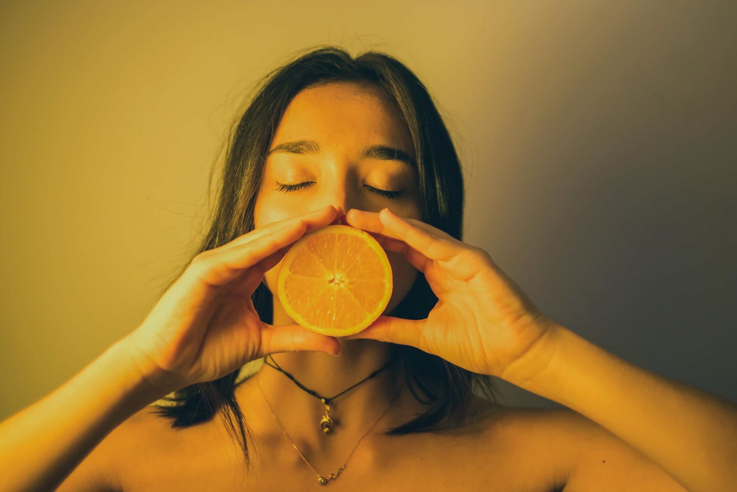 A person with long brown hair holds an orange slice over her mouth to represent satya, or truth. She is wearing a necklace above her collarbone.