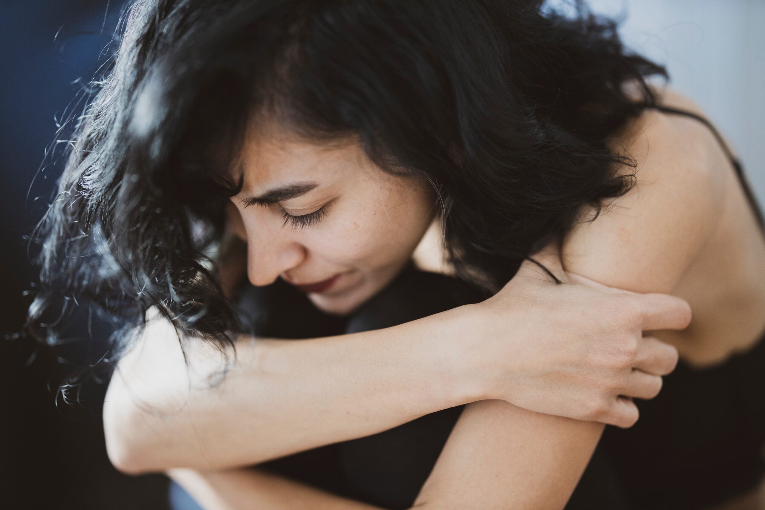 Person with black hair and red lips sits with arms crossed, bugging her knees. Her head is bowed toward her knees.