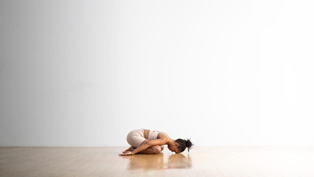A Black woman wearing cream colored tights and top practices Child's Pose (Balasana). She is on a wood floor against a white backdrop.