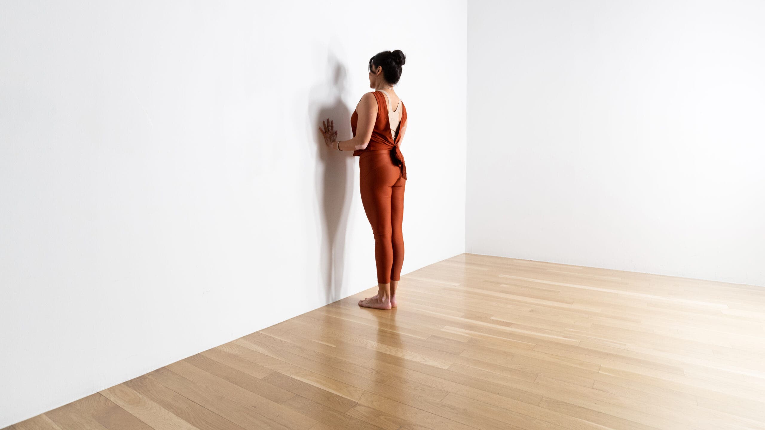 A woman with dark hair faces a wall with her hands pressing against it. 