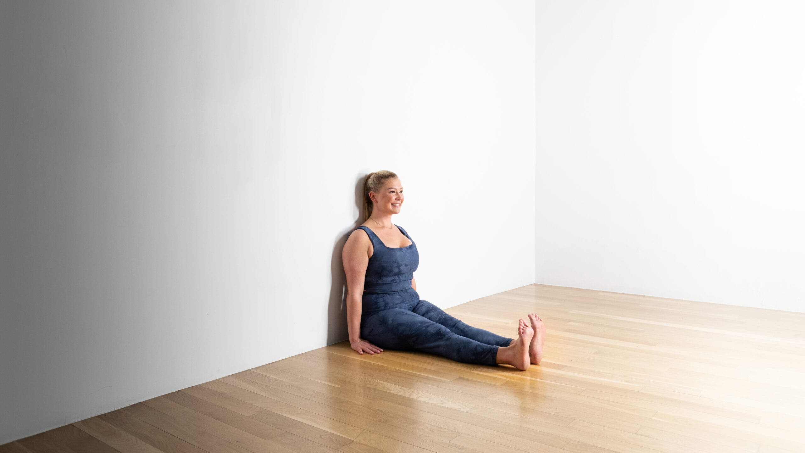 A woman with blonde hair sits with her back against a white wall, practicing Dandasana (Staff Pose)