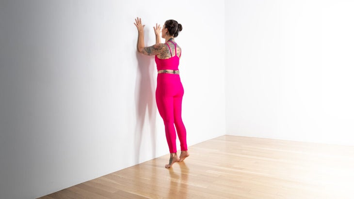 A woman in bright pink tights practices forearm plank against a white wall.