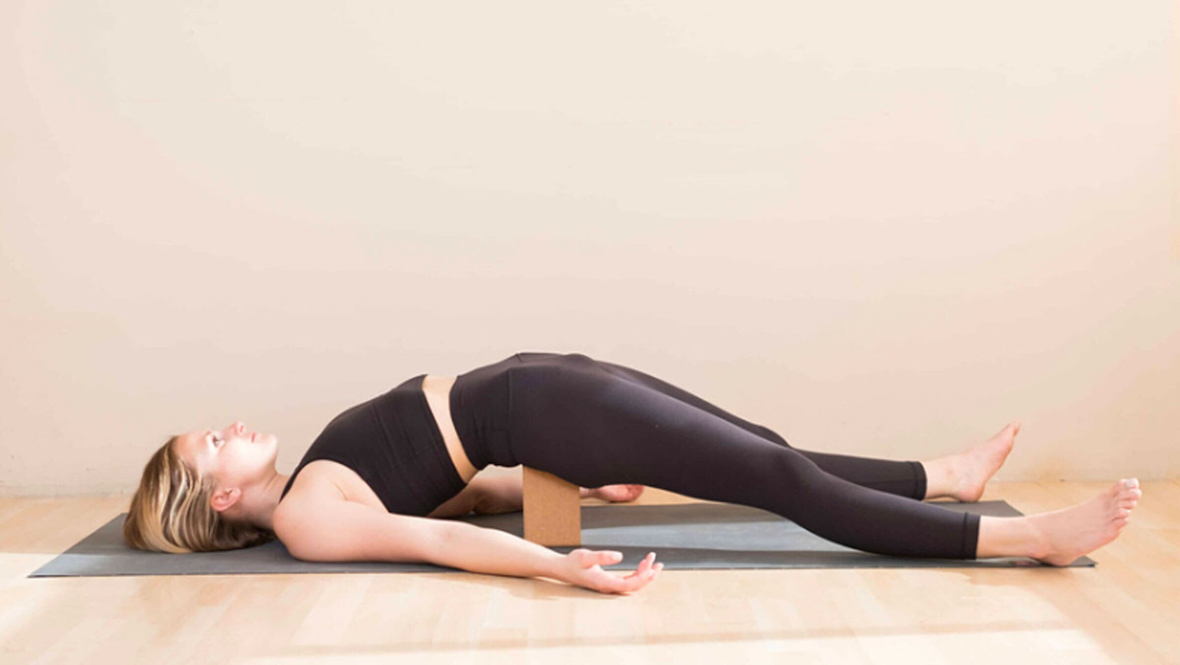 Woman lying on a yoga mat with a block beneath her sacrum to create a slightly backbend in her lower back. Her arms are resting alongside her body and her legs are extended straight from her hips.