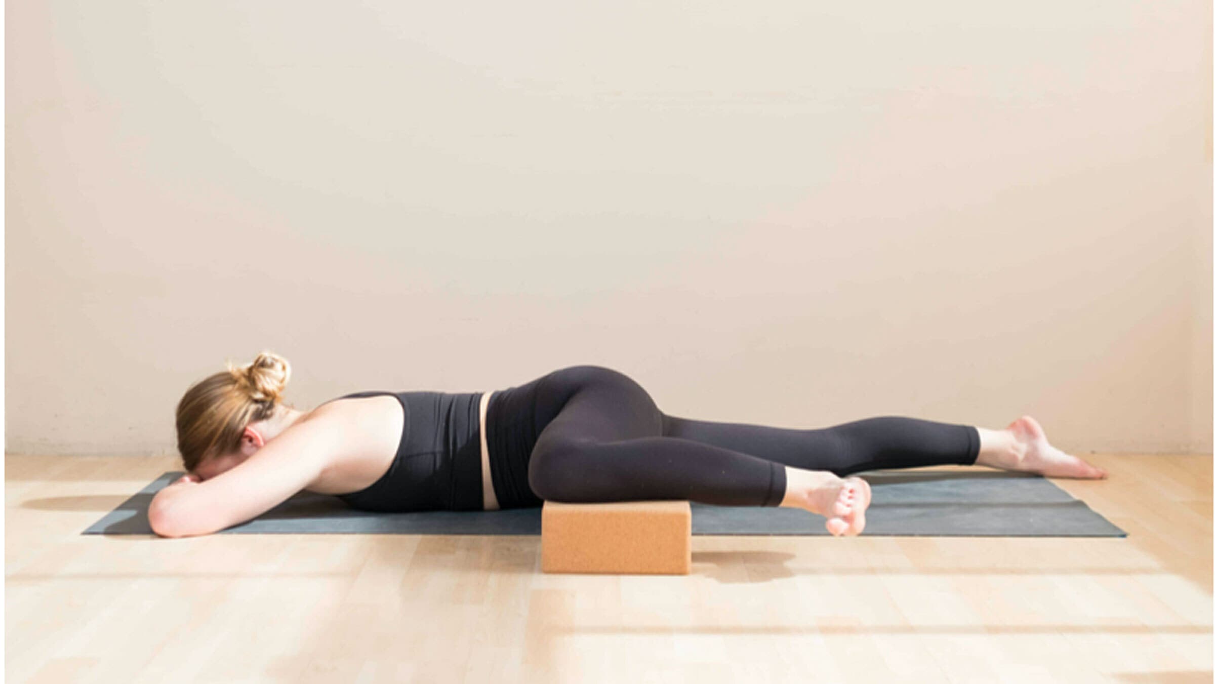Woman lying face-down on a yoga mat with her left knee bent. She's using a yoga block beneath her knee to stretch her adductor muscles.