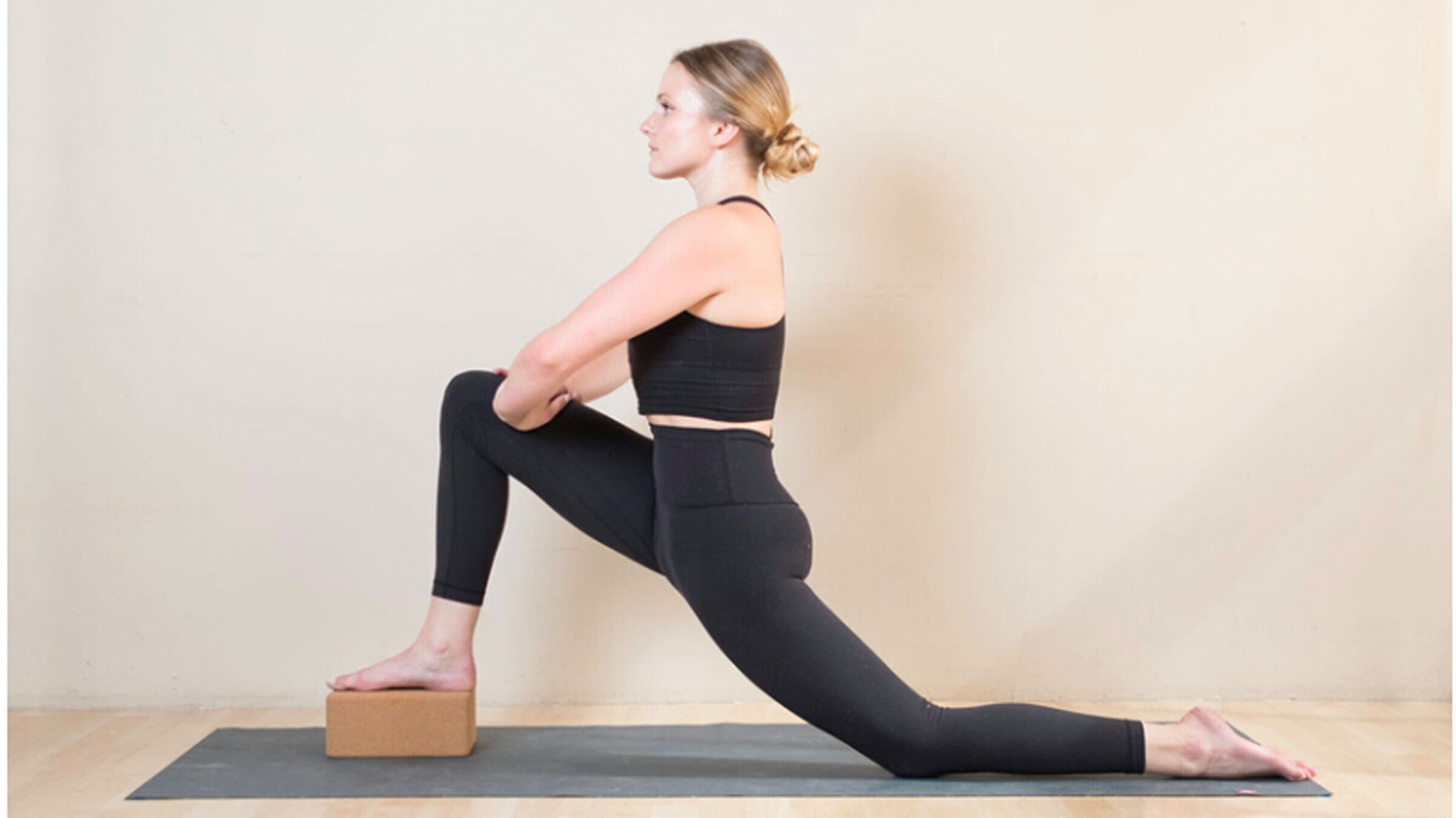 Woman in a lunge on a yoga mat with one knee on the mat and the other foot on a yoga block. Her hands are on her front thigh.