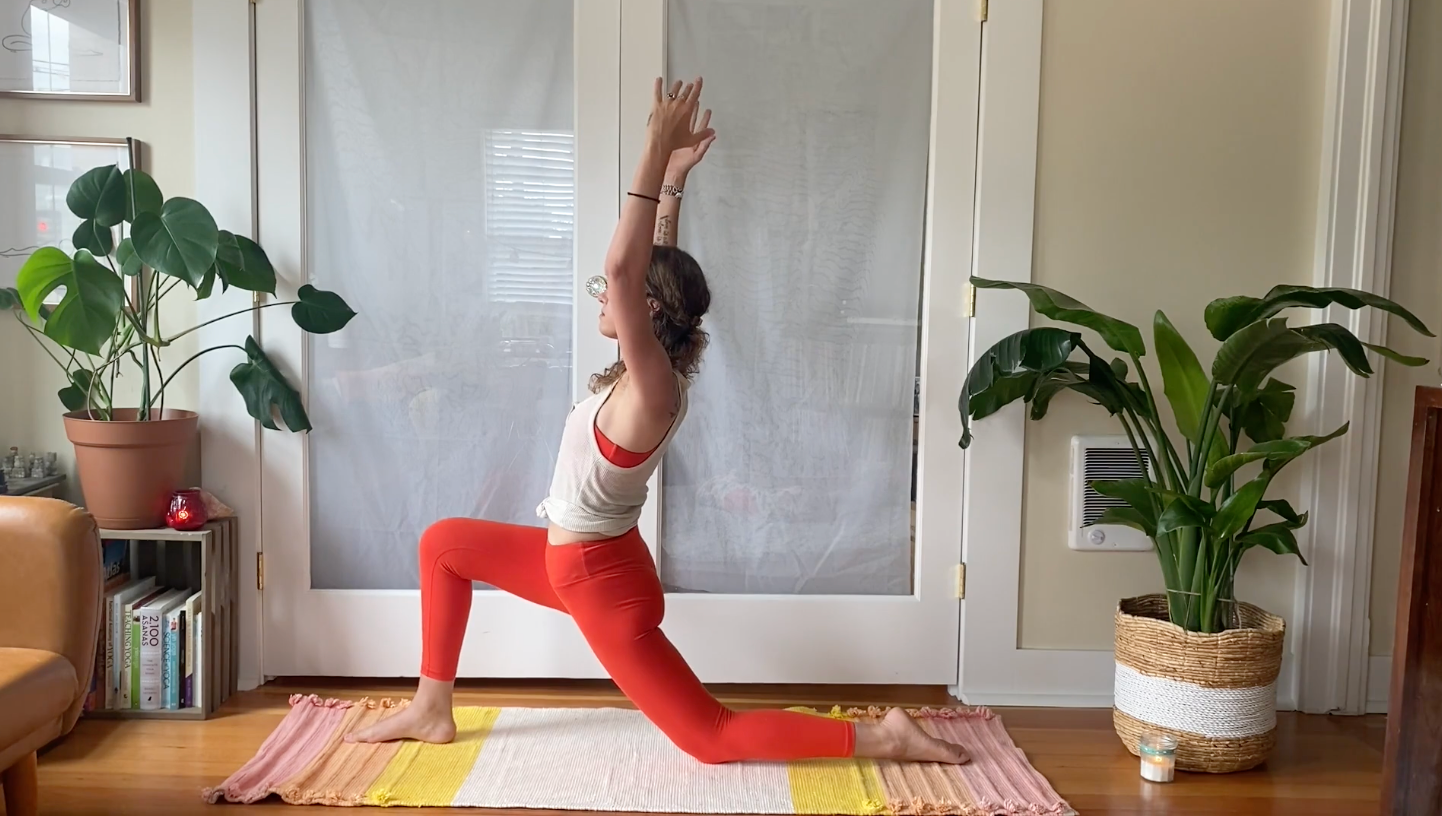 Woman on her yoga mat with one knee down and the other in a low lunge with her arms alongside her ears stretching her hips