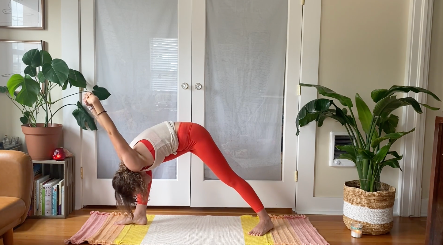 Woman standing on her yoga mat in a Warrior 1 stance but with her fingers interlaced behind her back as she leans forward at her hips