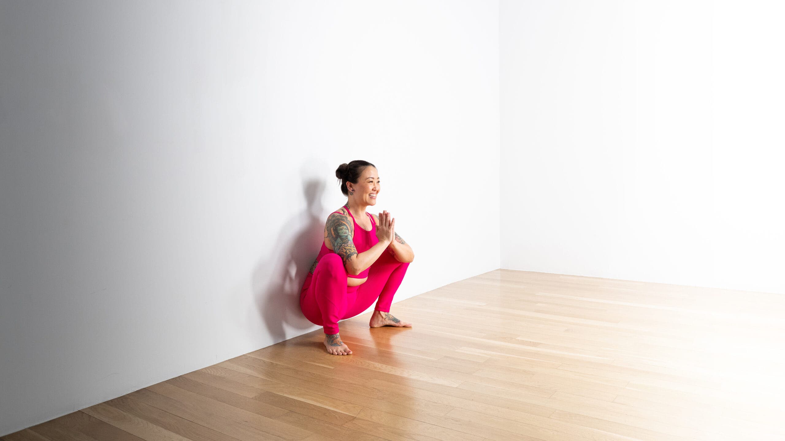 A woman with dark hair wears bright pink tights practices Malasana, Garland Pose. She is squatting with her hips against the wall.