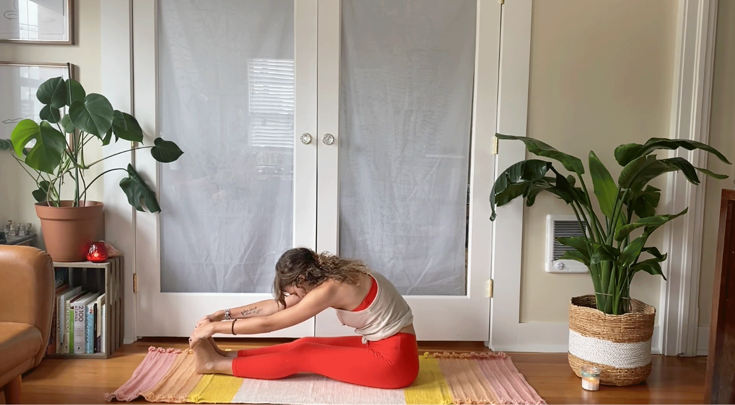 Woman seated on her yoga mat with her legs straight in front of her and her hands on her feet as she leans forward in Paschimottanasana