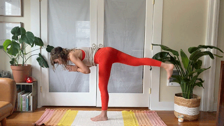 Woman balancing on one leg on her yoga mat with her right leg extended parallel to the floor and her hands at her heart in prayer