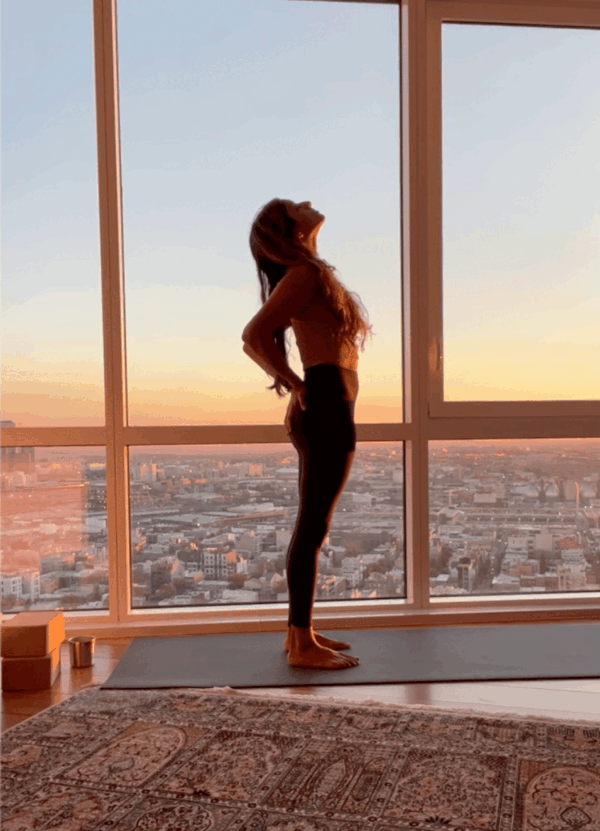 Woman standing on her yoga mat with the sunrise behind her practicing a gentle backbend