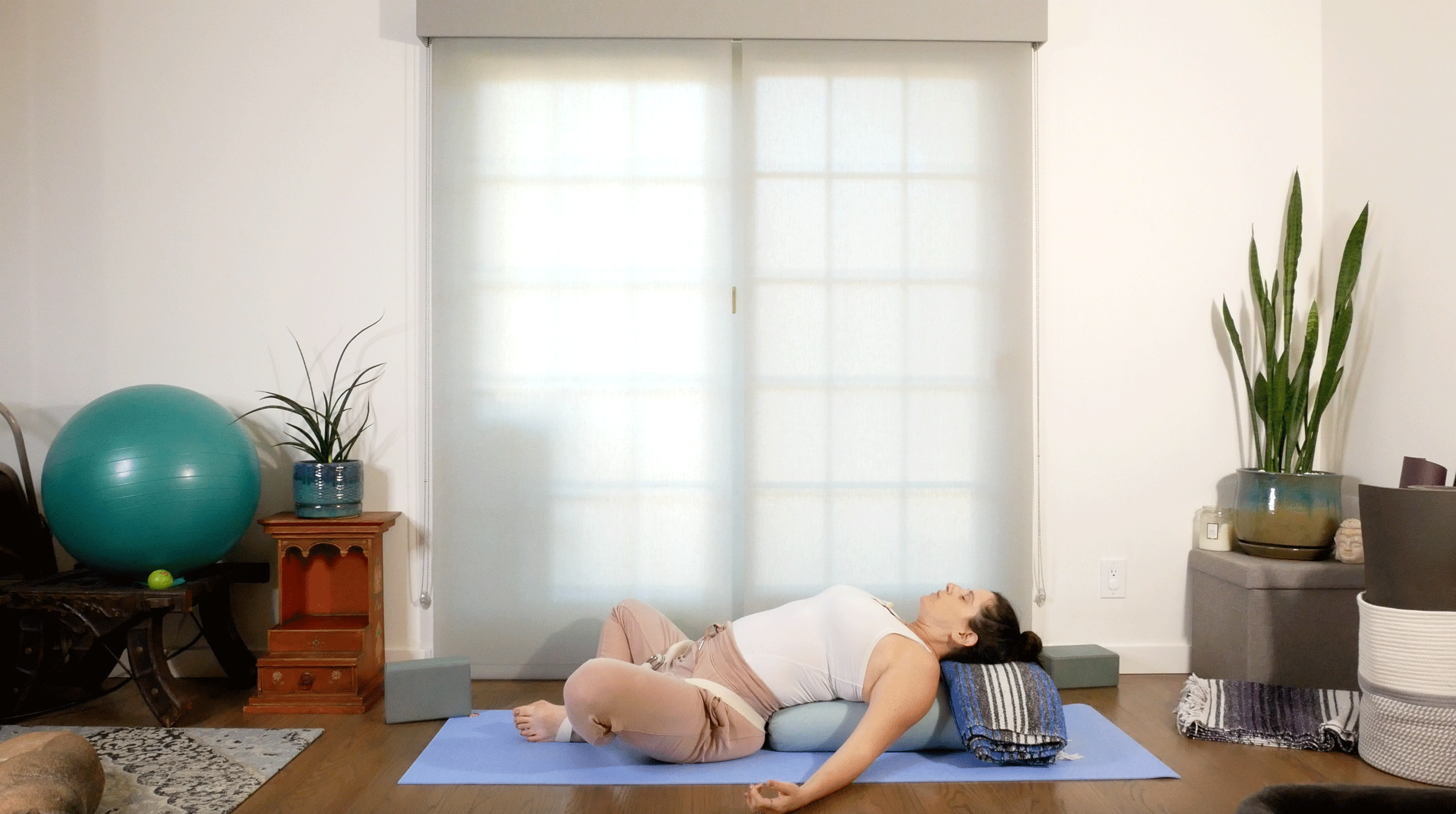Woman lying on her back on a bolster with her knees bent and her arms resting at her sides in a soothing supported yoga pose