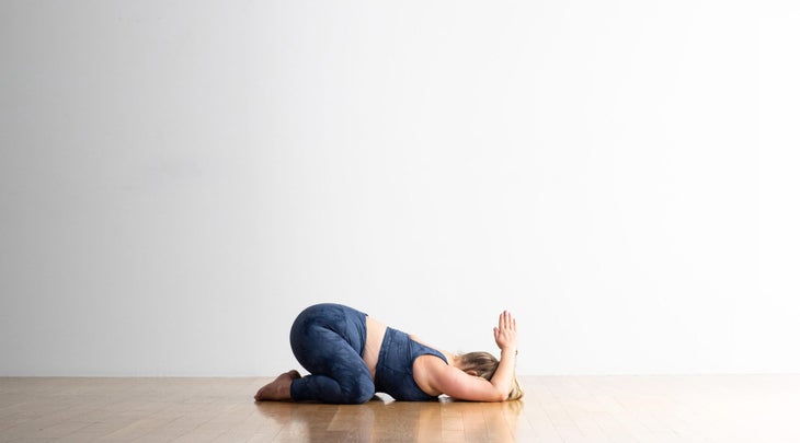 A woman with blond hair and blue clothing practices Child's Pose with hands behind the head in Anjali Mudra