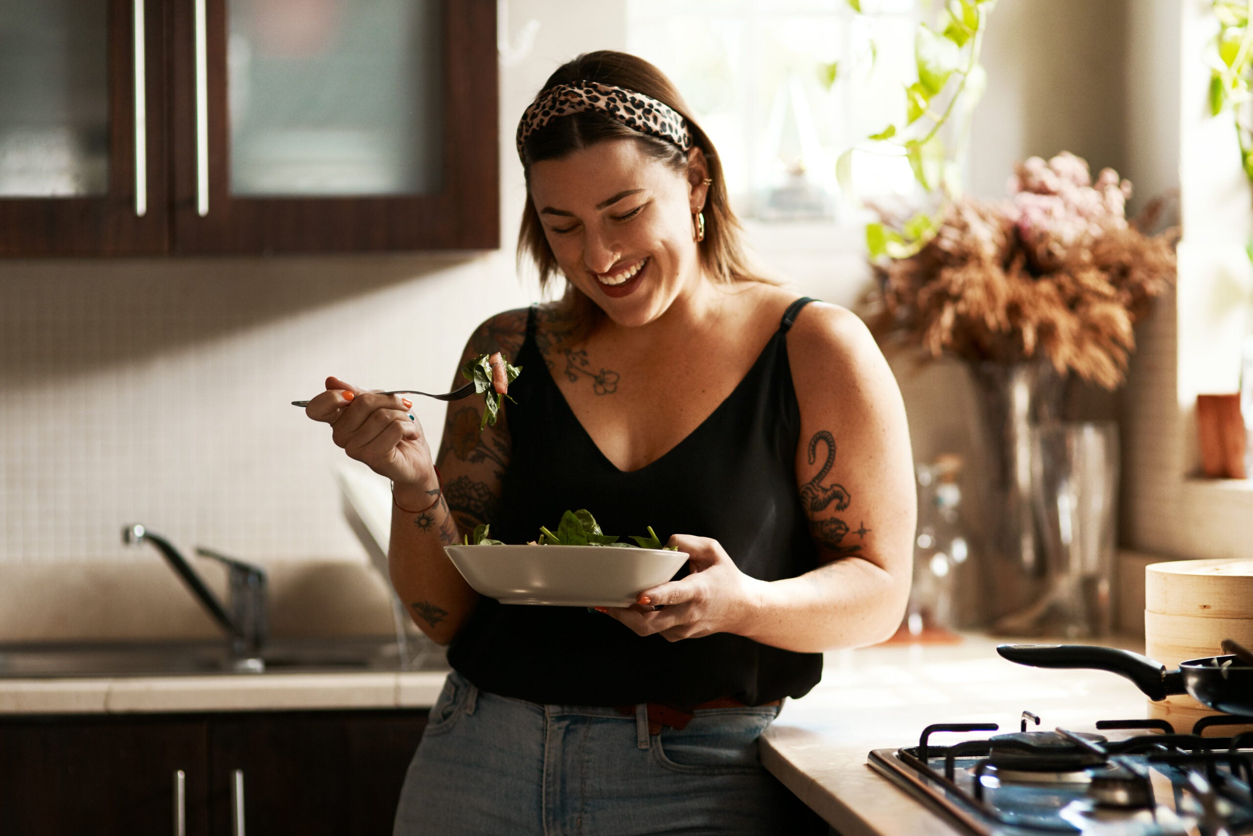 A mid-sized woman stands beside a kitchen counter holding a white bowl of food. She has blond hair and tattoos on her arms. She wears a black top and blue jeans. Brown cabinets and a plant are behind her.