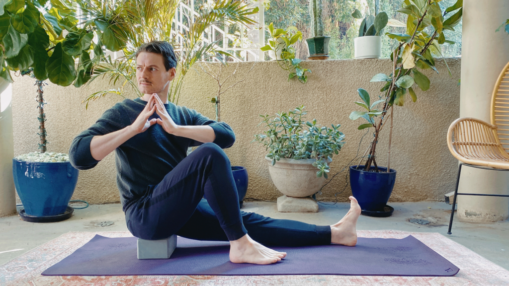 Man sitting on a block on a yoga mat with one knee bent and another leg straight. He is twisting to the right.