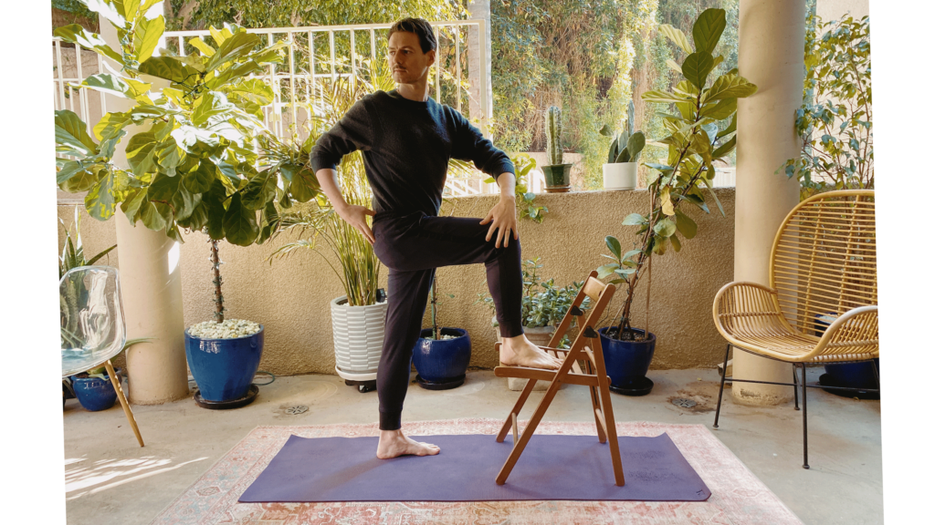 Man standing on a yoga mat with one foot on the seat of the chair and the upper body twisting to the right