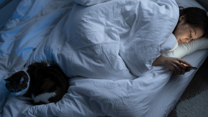 A woman curled up in bed under a blanket with her cat and her phone is so worried that she can't sleep