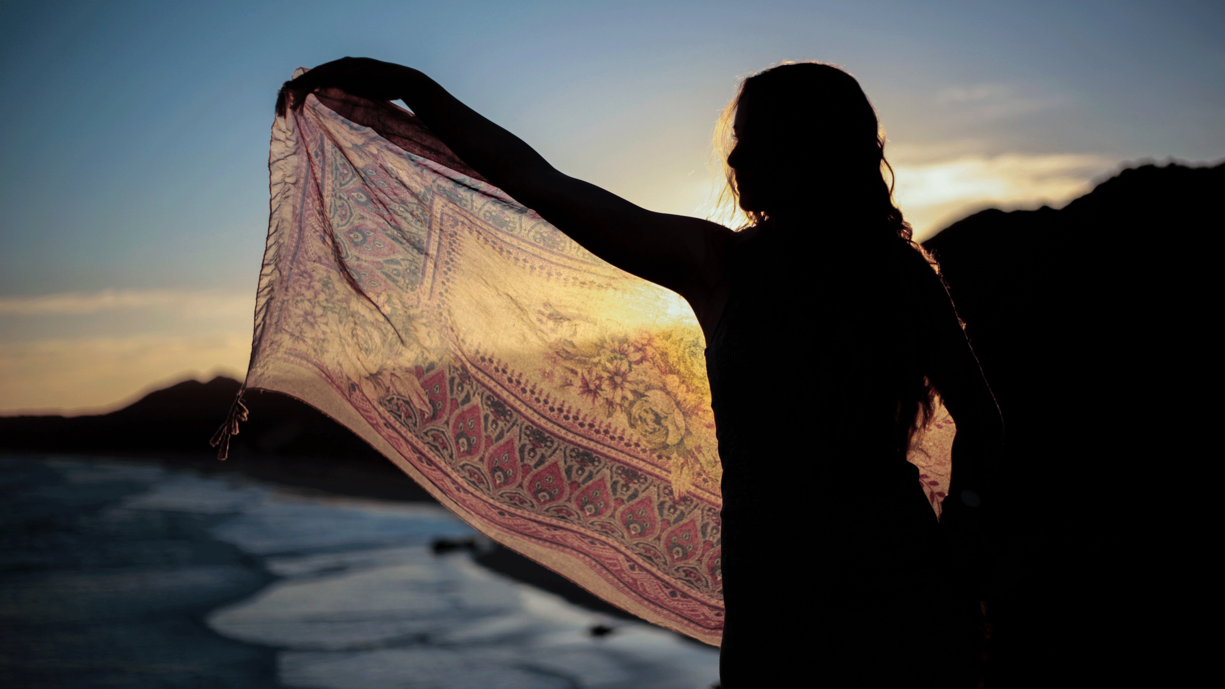 Woman standing on a cliff overlooking the ocean and holding her left arm out and draping a scarf through which you can see sunlight