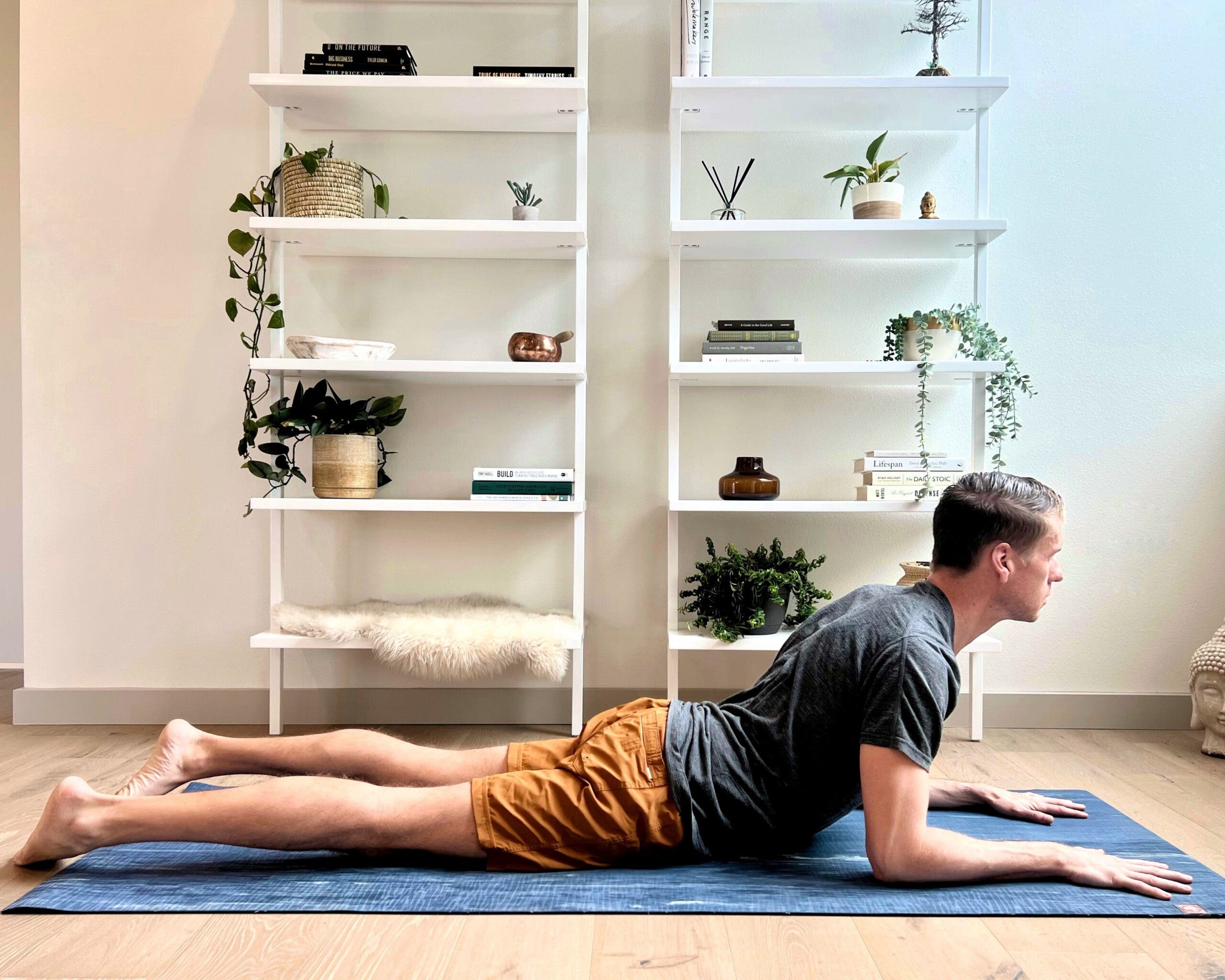 A man practices Sphinx pose in a white room with two white book cases behind him. He is wearing a gray T-shirt and orange shorts.