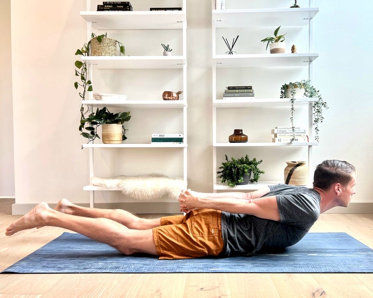 A man in a gray t-shirt and orange shorts practices Salabhasana (locust pose) in a white room with shelves behind him