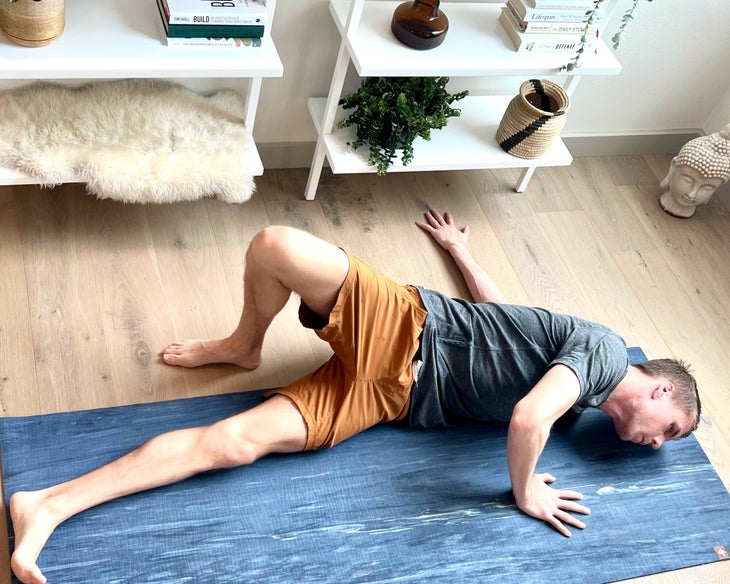 A man wearing a gray T-shirt and orange shorts practices a supine twist. He is lying on a gray mat. He has on a gray T-shirt and orange shorts. White shelves are behind him.
