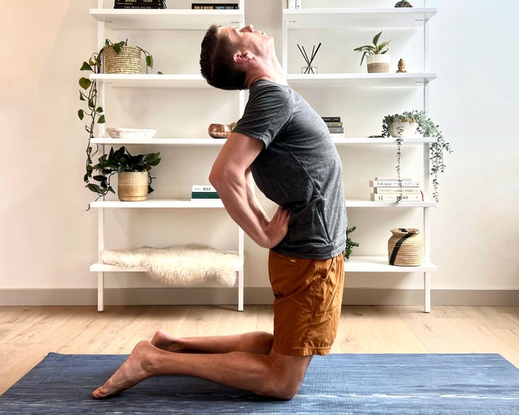 A man in a gray shirt and orange shorts practices Ustrasana (camel pose).  Behind him is a white wall and white shelves