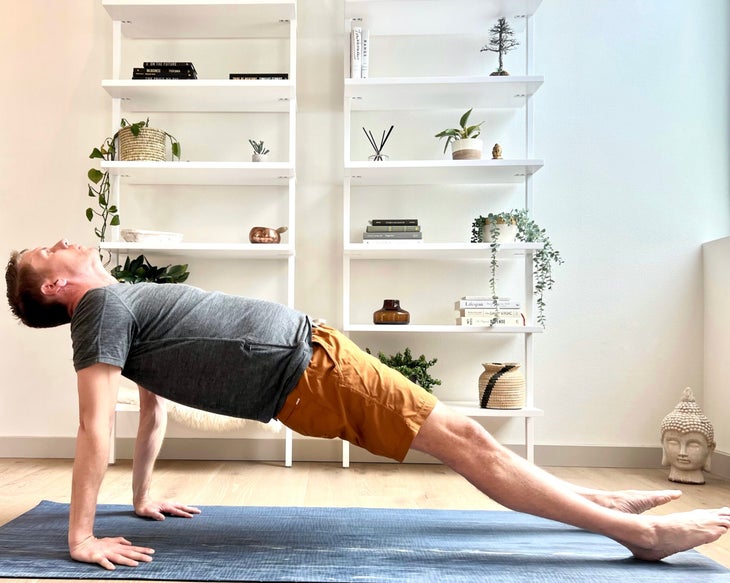 A man in a gray T-shirt and orange shorts practices Purvottanasana (Reverse or Upward Plank Pose) on a gray mat. A white wall and shelves are behind him