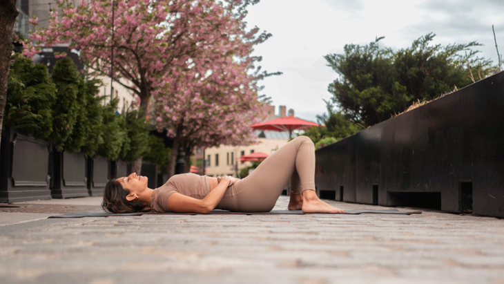 Woman lying on a yoga mat on her back with her knees bent and her feet on the mat. She's outside on a brick sidewalk with blossoming trees.