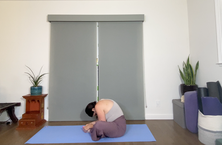 A woman with dark hair practices Baddha Konasana with a forward bend. She is wearing purple pants and a gray tank, sitting on a blue mat