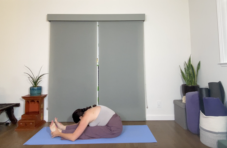 Woman folds forward in Pascimottanasana. She is sitting on a blue rug. She is wearing loose purple pants and a gray T-shirt