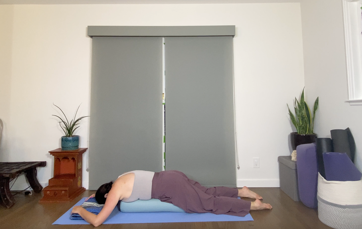 A woman practices a prone Savasana, lying over a blue bolster.