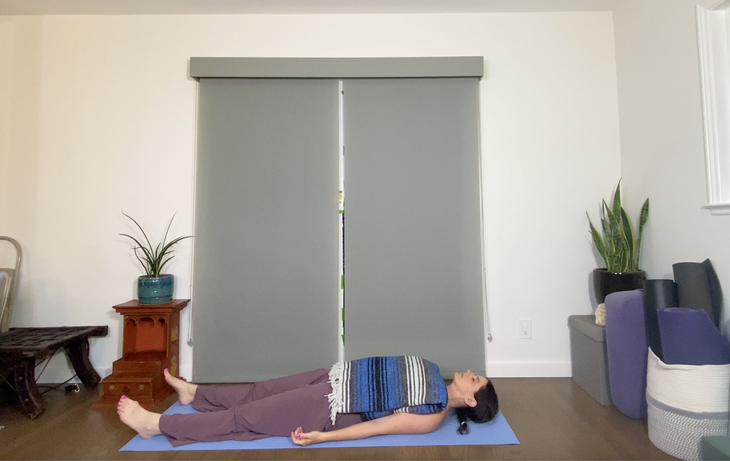 A woman lies in Savasana, corpse pose, with a blue striped blanket covering her torso