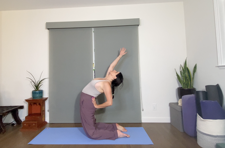 A dark-haired woman practices Camel Pose with one arm raised and the other hand on her hip