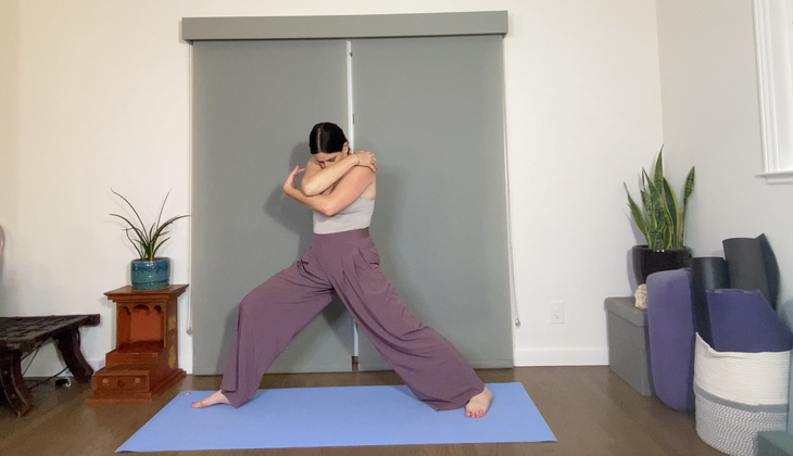 A woman with dark hair practices Warrior 2 Pose with her arms wrapped around to hug her body.