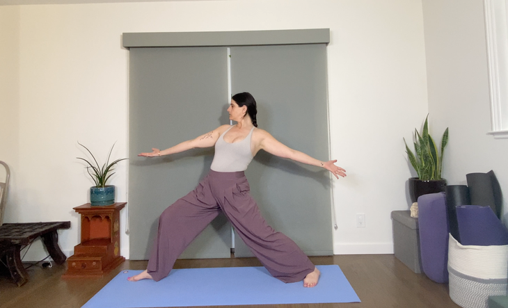 A dark-haired woman in loose purple pants practices a moving version of the Warrior 2 pose