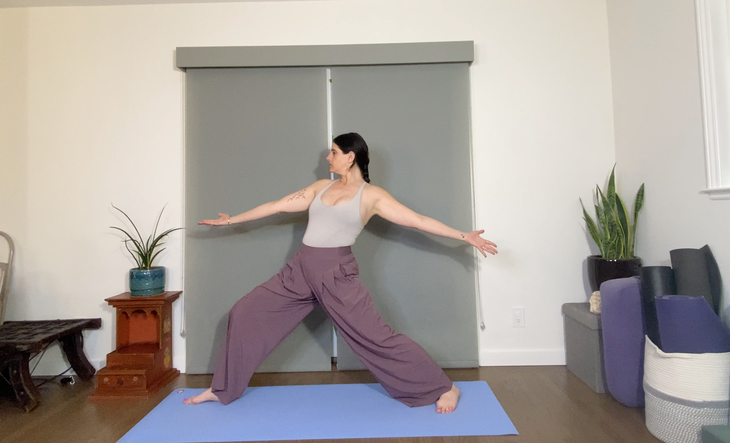 A dark-haired woman wearing loose purple pants practices a moving variation of Warrior 2 Pose