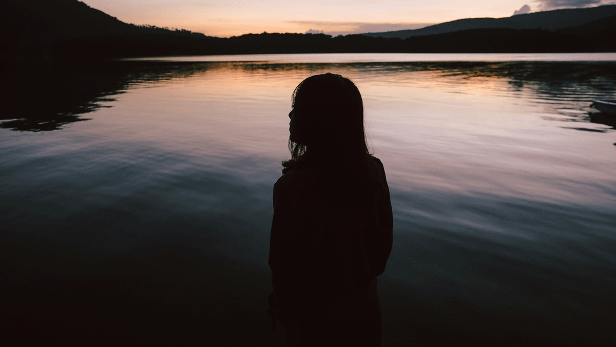 Woman standing at the edge of a lake surrounding by mountains at sundown contemplating her weekly astrology forecast