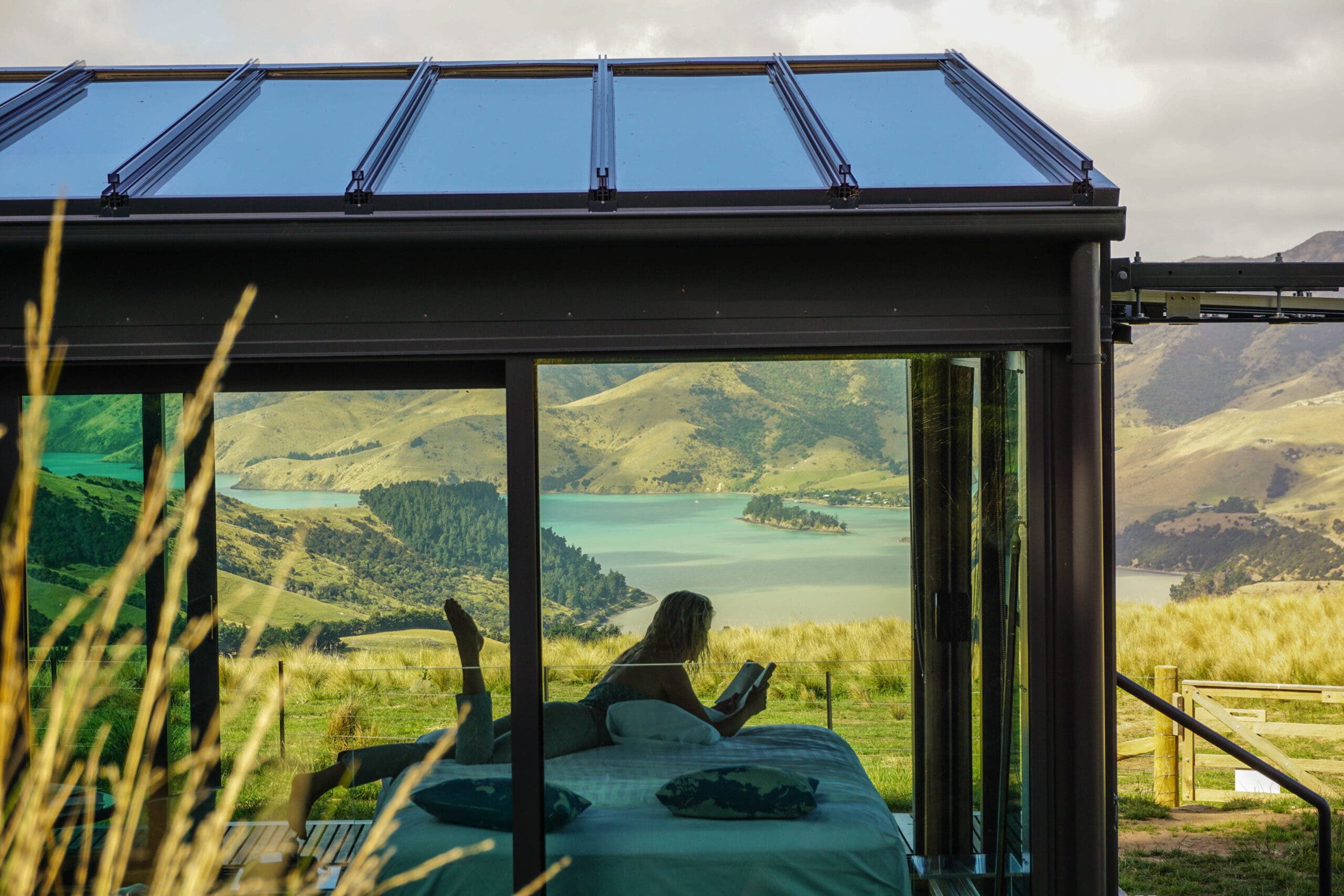 A person lying on a bed reading a book in a glass walled structure. Beyond the glass cabin is a view of hills, mountains, and a body of water in New Zealand