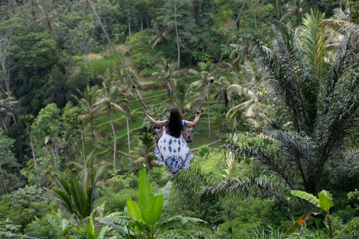 A woman in a white dress with blue flowers swings over the jungle in Bali.