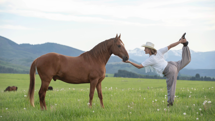 A woman practices a yoga dancer pose in a field in Montana while stroking a horse.  Behind her are mountains and a cloudy sky.