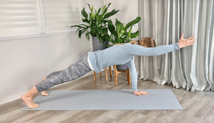 A woman practices a plank pose with one arm raised from the yoga mat
