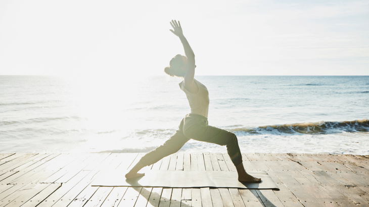 A woman practices yoga outdoors on a wooden deck near the ocean at sunrise