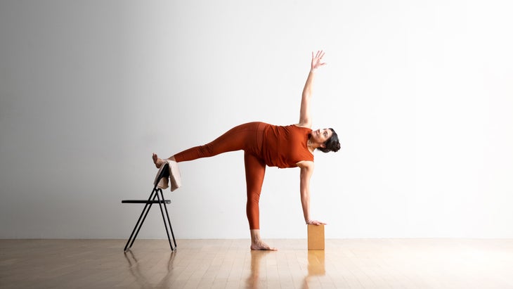 Noemi Nunez practices Half Moon Pose (Ardha Chandrasana) with her foot on a metal folding chair.  She has black hair and wears copper colored clothes.  She is on a light wooden floor with a white wall in the background.