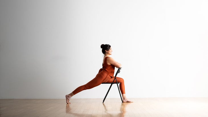Noemi Nunez practices a High Lunge while straddling a metal folding chair.  She has black hair and wears copper colored clothes.  She is on a light wooden floor against a white wall in the background.