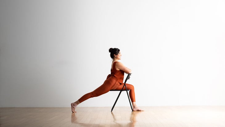 Noemi Nunez practices a High Lunge by straddling a metal folding chair. She has dark hair and is wearing copper colored clothes. She is on a light wood floor against a white wall in the background.