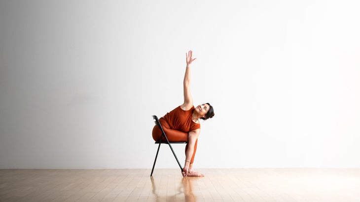 Noemi Nunez practices chair pose with a twist to the right side.  by straddling a metal folding chair.  She has black hair and wears copper colored clothes.  She is on a light wooden floor against a white wall in the background.