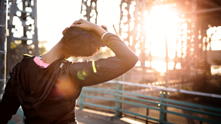 Woman runner practicing a neck and shoulder stretch at sunrise beneath a bridge