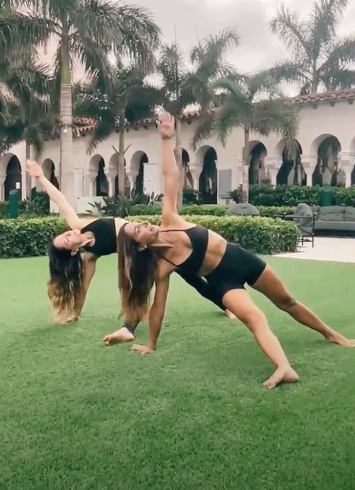 Two yoga teachers practicing Fallen Triangle on a grassy lawn with palm trees in the background