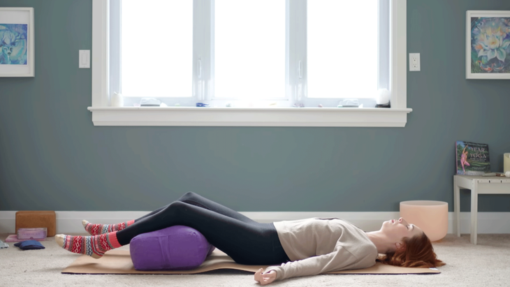 Yoga with Kassandra in Savasana on her yoga mat with a bolster beneath her knees