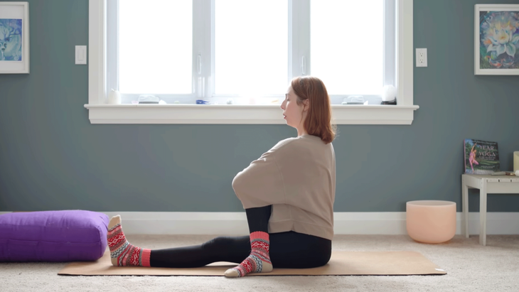 Yoga with Kassandra sitting on her yoga mat sitting upright with her right knee pulled into her chest and her right foot alongside her left knee